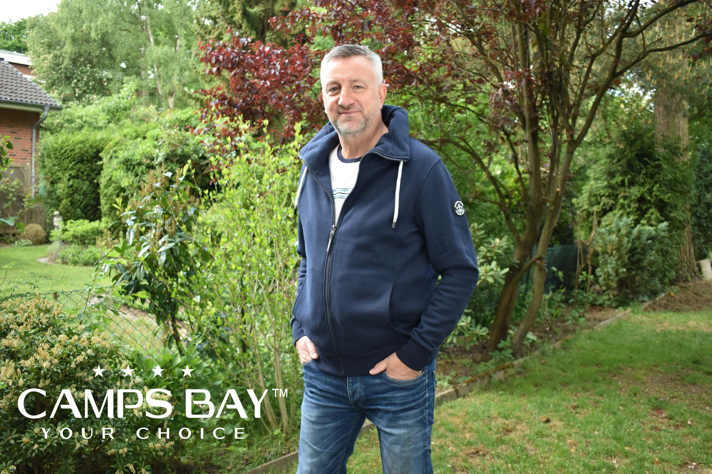 Mann mittleren Alters trägt eine blaue Camps Bay Sweatjacke und steht vor einer Garten-Kulisse