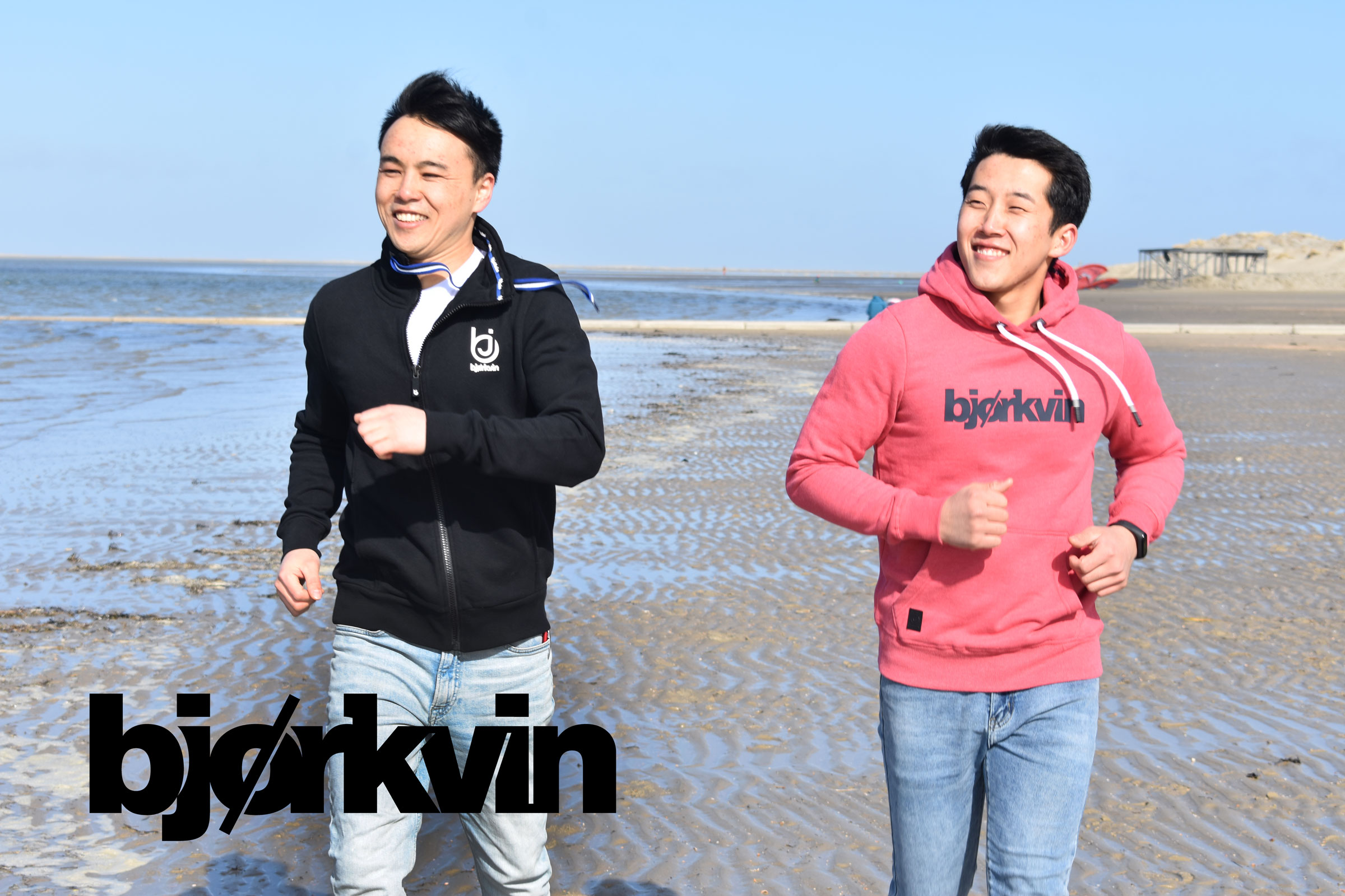 Zwei junge Männer in Björkvin Sweatshirts joggen nebeneinander am Strand